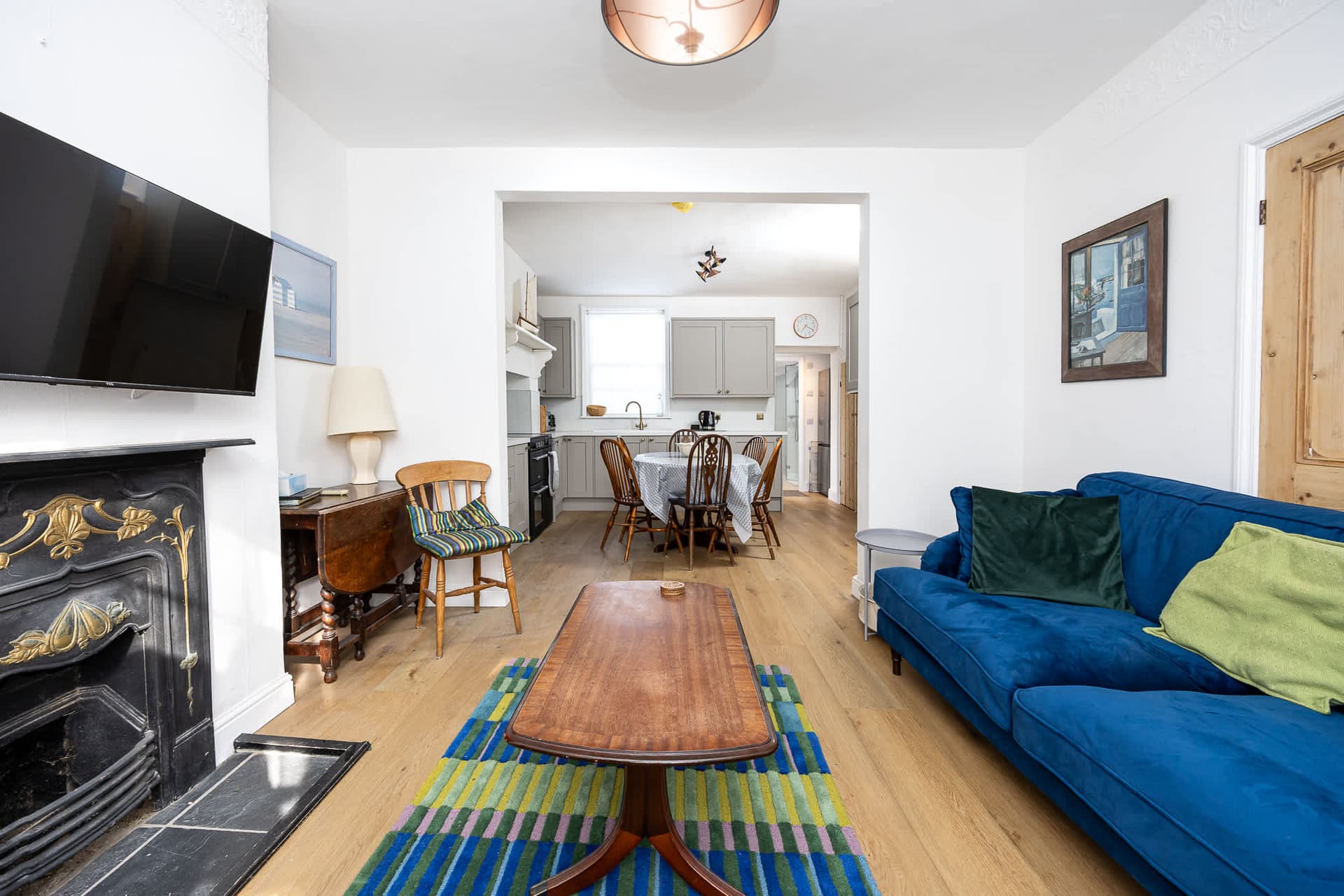 Relaxed downstairs living space with natural light, wooden floors and modern, thoughtful furnishings. A classic fireplace adds character, with family dining and a modern kitchen beyond. Ideal for unwinding or giving guests extra space.