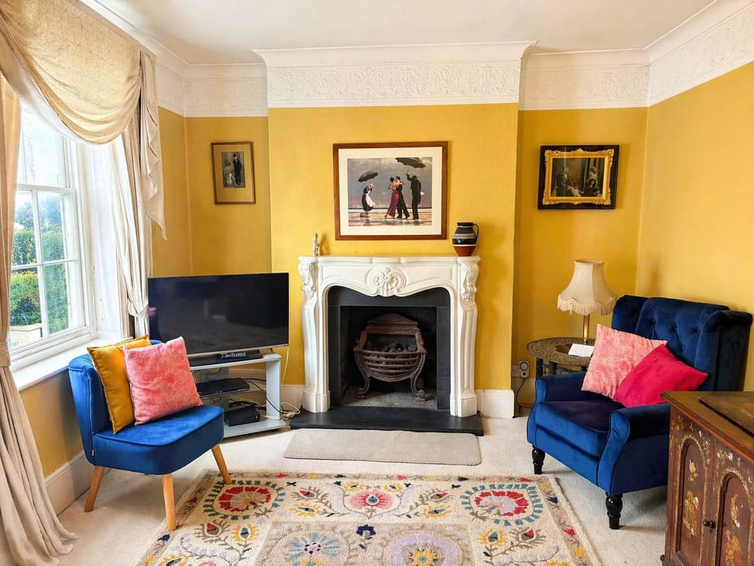 Characterful period sitting room with mustard walls, blue chairs and an ornate fireplace
